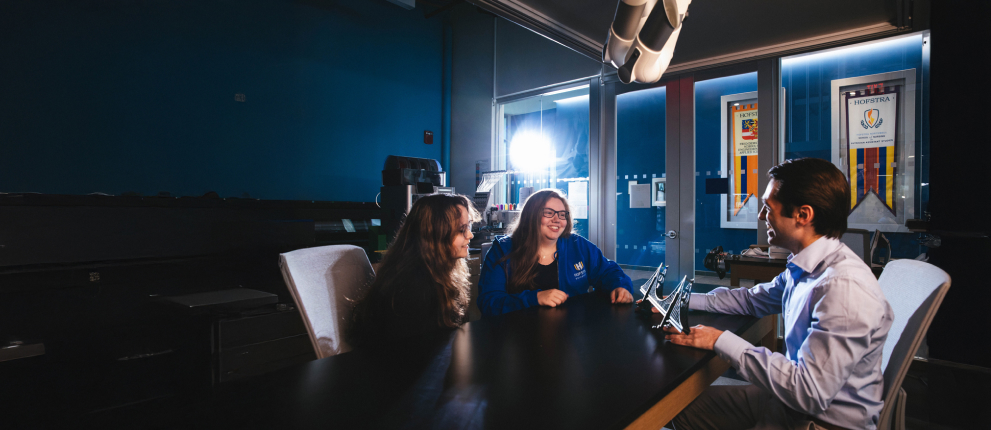 Three people sit around a dark conference table in a modern office, engaged in conversation. One person holds a tablet, while sunlight streams through large windows behind them, illuminating banners outside.