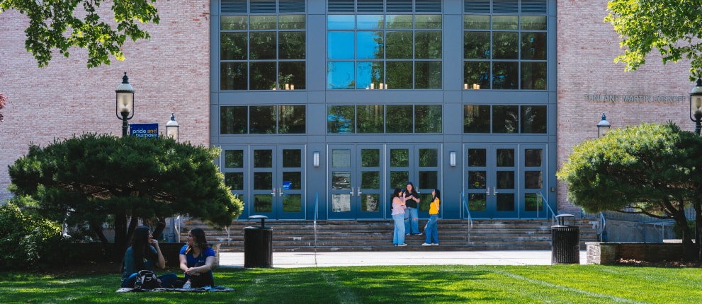 Students sit and talk on a grassy lawn and others stand near the entrance of a large building with many windows and doors on a sunny day.