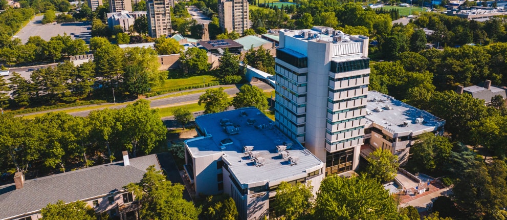 Aerial view of a modern multi-story building surrounded by dense green trees, nearby roads, and other buildings under a clear sky.