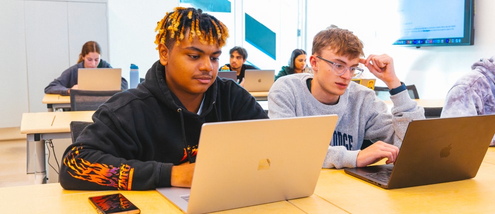 Two students sit side by side at a classroom desk, working on laptops. Other students are also working at desks in the background. A phone and water bottle are visible on desks. Classroom is bright and modern.