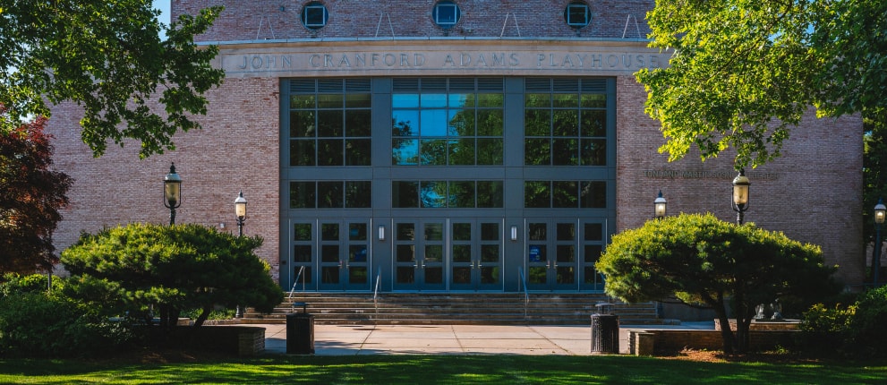 The entrance of John Cranford Adams Playhouse, a brick building with large glass windows and steps, surrounded by green trees and neatly trimmed bushes.