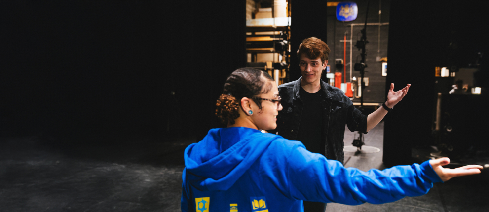 Two people stand on a stage; one wearing a blue hoodie gestures with an outstretched arm, while the other, in black, faces them with open arms, with stage equipment and a dimly lit background visible.
