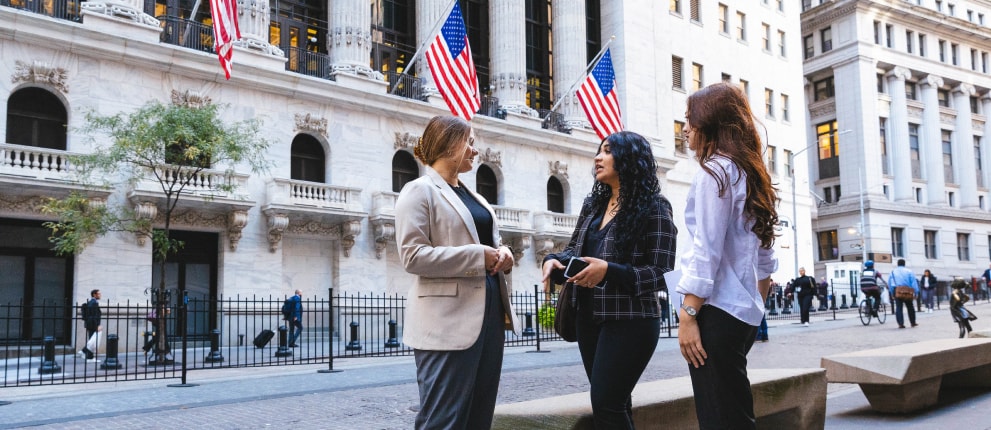 Three women in business attire stand talking outside a historic building with large columns and American flags, likely on Wall Street. Other people walk nearby, and the scene is set in an urban environment.