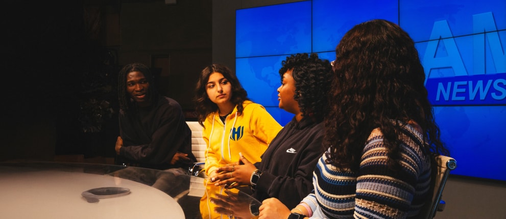Four people sit at a news desk having a discussion; behind them is a large digital screen with a blue news graphic. They appear engaged and focused on the conversation.