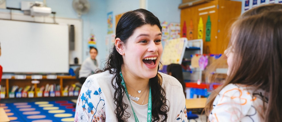 A woman with long dark hair smiles widely while talking to a young girl in a colorful, cheerful classroom with bookshelves and educational posters in the background.