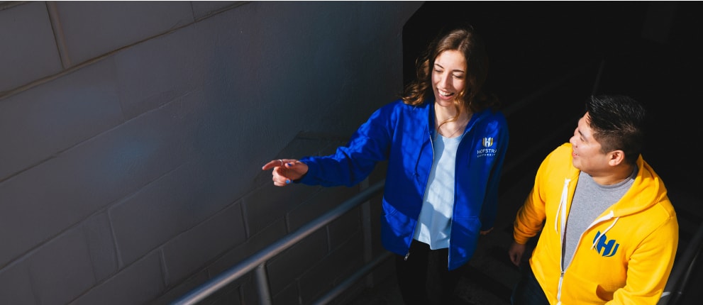 Two people in branded jackets walk up a staircase. The woman in a blue jacket points ahead while smiling, and the man in a yellow jacket looks at her, both appearing cheerful and engaged in conversation.