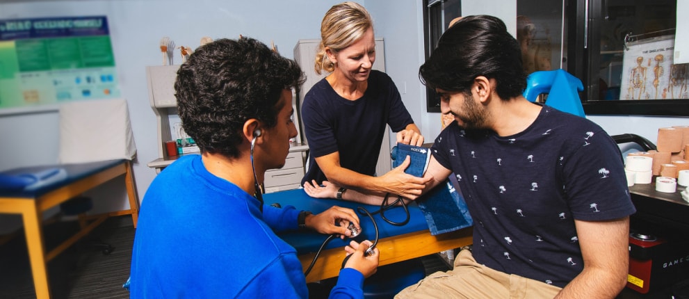 A healthcare professional helps a young man have his blood pressure checked by another young man using a blood pressure cuff and stethoscope in a clinical setting.