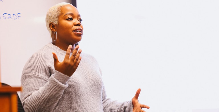 A person with short blond hair and hoop earrings speaks while gesturing with their hands, standing in front of a whiteboard in a classroom or meeting setting.