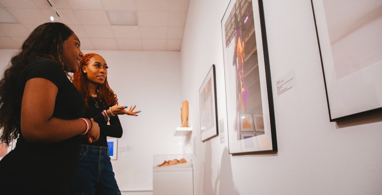Two women stand in an art gallery, closely observing and discussing a framed artwork on the wall. The gallery is bright and features other sculptures and framed art pieces.