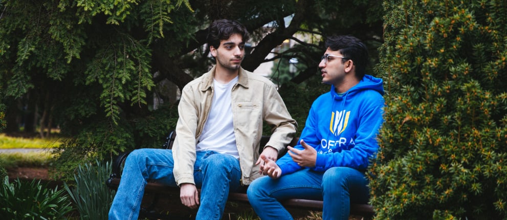 Two young men sit on a bench outdoors, surrounded by green trees. One wears a beige jacket and jeans, while the other wears a blue hoodie and glasses. They appear to be having a serious conversation.