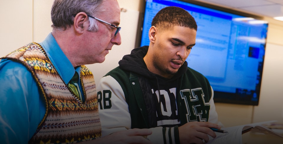 An older man in glasses and a patterned vest helps a young man wearing a varsity jacket with his work in a classroom, with a large monitor displaying a computer screen in the background.