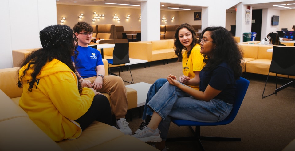 Four students sit in a lounge area, engaged in conversation. Two wear yellow shirts, one wears a blue shirt, and one wears a yellow hoodie. They are seated on modern couches and chairs in a brightly lit room.