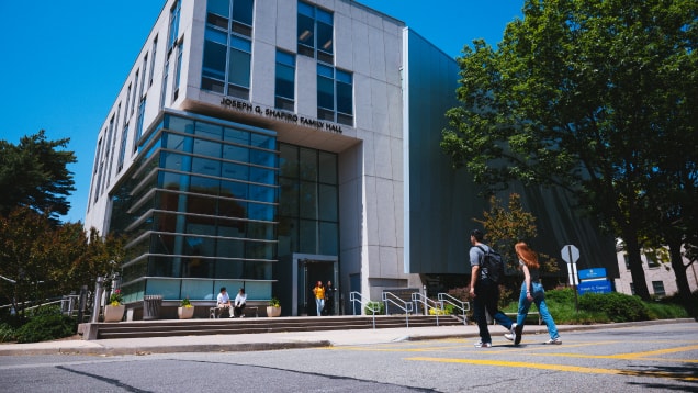 Two people walk across a campus road toward Joseph G. Shapiro Family Hall, a modern building with large glass windows. Several people sit on steps outside under a sunny, blue sky, surrounded by trees.
