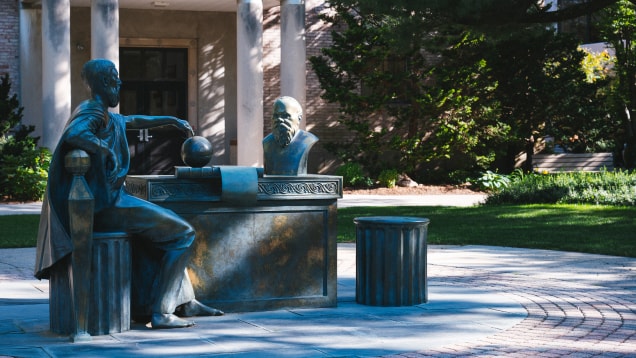 A bronze outdoor sculpture features a seated figure pointing at a globe on a desk, facing a bust, with columns and greenery in the background, on a university campus.