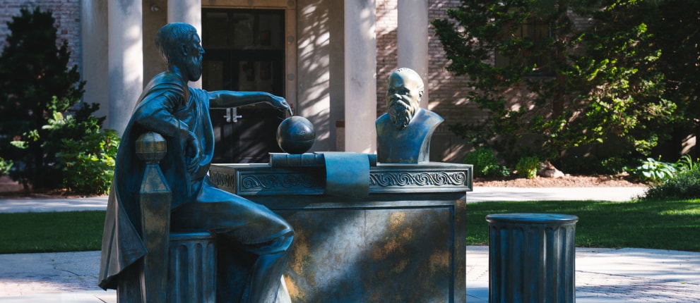 A bronze outdoor sculpture of two scholars sits at a table with scrolls and a globe, one seated and leaning back, the other resting his head on his hand, with trees and a building in the background.