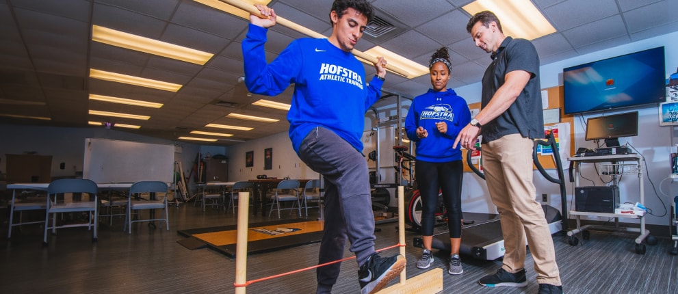 A student in a blue Hofstra Athletics shirt balances on one leg on a board, holding a wooden bar, while another student and an instructor supervise in a classroom with exercise equipment.