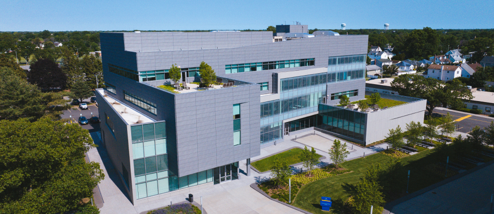 A modern, multi-story building with large windows and rooftop gardens, surrounded by trees and residential houses, under a clear blue sky.