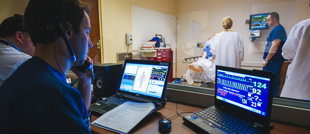 A person monitors two laptops displaying medical data in a control room, while several healthcare professionals attend to a patient in a hospital room visible through a window.