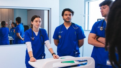 Three medical students in blue scrubs with stethoscopes stand around a hospital cart in a bright room, appearing attentive and engaged in discussion.