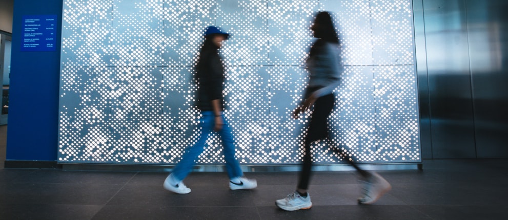 Silhouettes of students walking past a glowing geometric light wall at Hofstra University