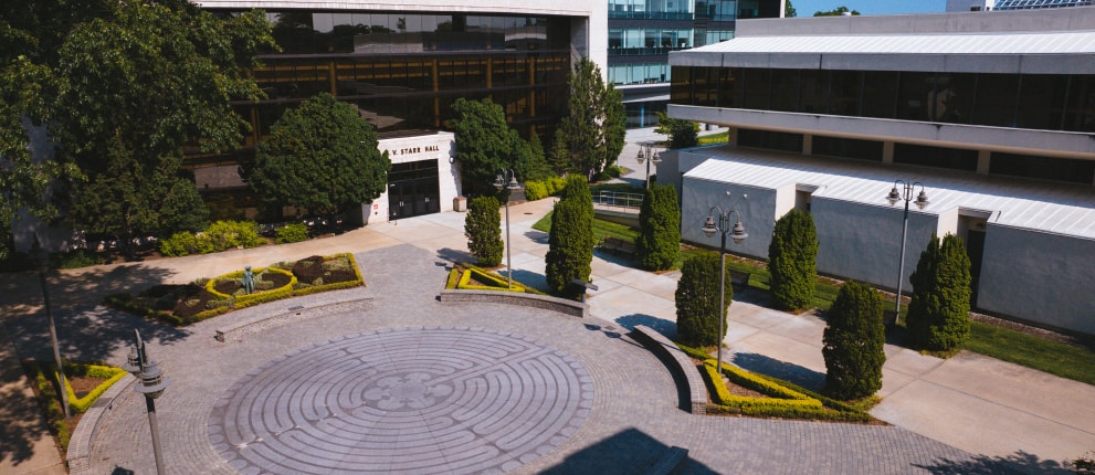 A circular stone labyrinth is surrounded by landscaped greenery and trees between modern campus buildings under a clear sky.