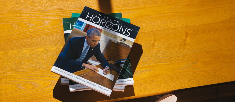 A stack of Hofstra Horizons magazines featuring a cover photo of Barack Obama in a suit writing at a desk, placed on a wooden surface with sunlight casting shadows.
