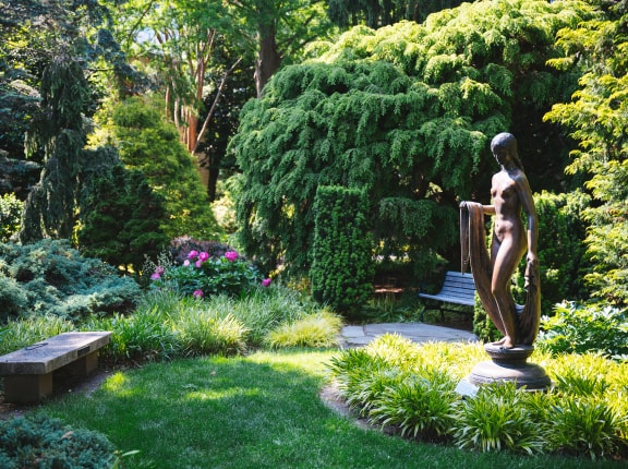 A lush garden with green trees and bushes, a stone path, two benches, pink flowers, and a bronze statue of a woman holding fabric, surrounded by well-manicured grass. Sunlight filters through the foliage.