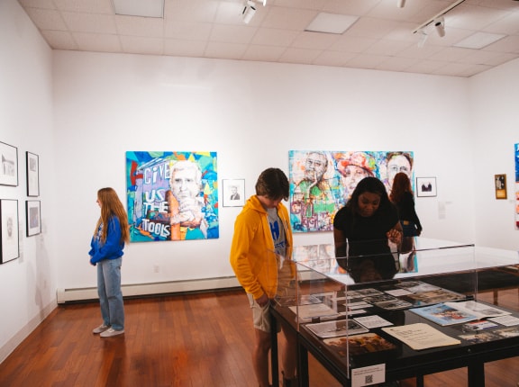 Four people view artwork and displays in a brightly lit art gallery with white walls, wooden floor, and colorful paintings and photos hanging and displayed in cases.