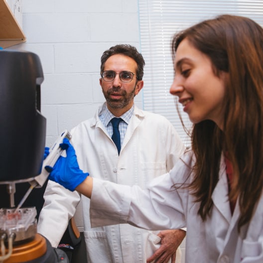Two scientists in lab coats work in a laboratory. The woman in the foreground operates equipment with a pipette, while the man in glasses and a tie observes her closely.